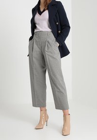 Marineblauer Mantel, hellrosa V-Ausschnitt-Pullover, hochtaillierte karierte Hose und beigefarbene Lack-Stiefeletten. Die Hose hat Falten und Taschen.