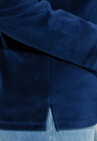 Top de manga larga de terciopelo suave en azul marino, con un dobladillo dividido; cuenta con una tela texturizada y mangas ajustadas, superpuesto a unos vaqueros azul claro.