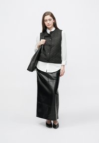 Črna prešita brezrokavnik čez belo srajco z gumbi, v kombinaciji s črno usnjeno maxi krilo in salonarji, drži črno torbo za na ramo.