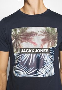 Osoba v námořnickém modrém tričku s grafickým vzorem palmových stromů a tropických listů, a s textem "JACK & JONES" tučným bílým písmem.