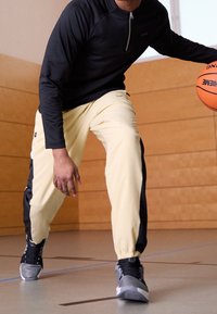 Schwarzer Reißverschluss-Oberteil, gelbe Sporthose mit schwarzen Seitenstreifen und graue Turnschuhe in einem Fitnessstudio mit Holzverkleidungen.