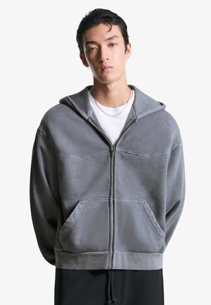 Graue Zip-Up-Hoodie mit Kapuze zum Zugbinden, ausgestattet mit einer Fronttasche und strukturiertem Stoff, getragen über einem weißen T-Shirt, auf schlichtem Hintergrund.