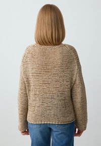 Beige Strickpullover mit lockerer Passform und überschnittenen Schultern. Verfügt über ein strukturiertes, offenes Gewebe und gerippte Bündchen. Kombiniert mit blauen Jeans.