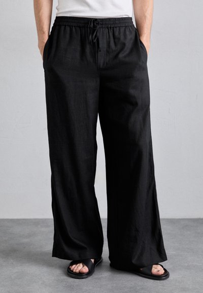 Schwarze weite Hosen aus leichtem Stoff mit einem elastischen Bund und einem Kordelzug. Getragen mit schwarzen Sandalen.