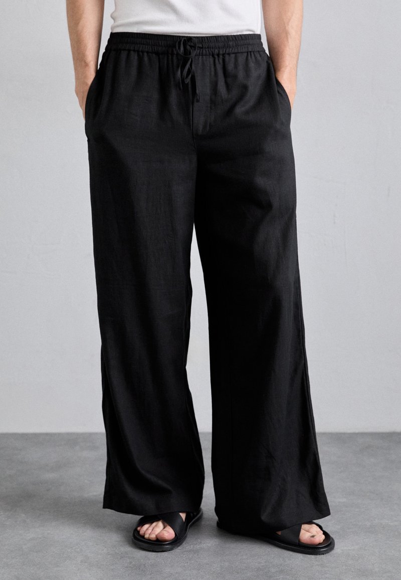 Pantalones negros de pierna ancha hechos de tela ligera con cintura elástica y detalle de cordón. Combinados con sandalias negras.