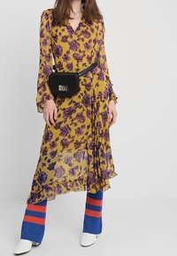 Žluté květinové šaty s fialovými vzory, dlouhé rukávy, volánkový lem; kombinované s modrými pruhovanými kalhotami a černou páskovou taškou. Bílé kotníkové boty.