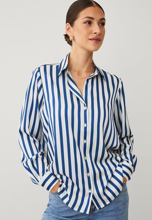 Woman wearing a blue and white vertical striped button-up shirt, with hands in pockets and gold earrings, looking right.
