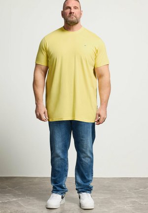 Man met kort haar en baard, gekleed in een geel T-shirt, blauwe spijkerbroek en witte sneakers, staand op een marmeren vloer tegen een witte muur.