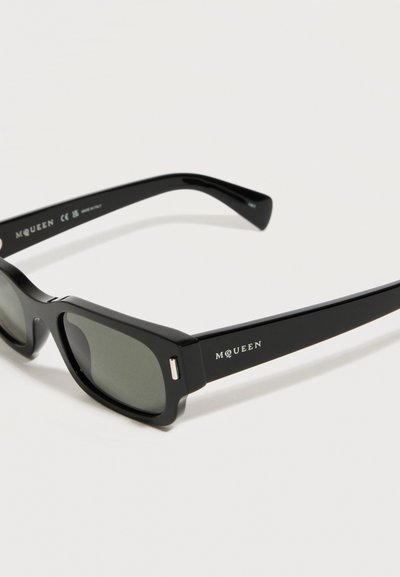 Μαύρα ορθογώνια γυαλιά ηλίου με σκούρους φακούς και λογότυπο "McQueen" σε λευκό χρώμα στο πλάι του σκελετού, εμφανιζόμενα πάνω σε λευκό φόντο.