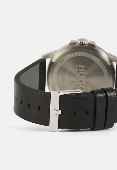 Bracelet de montre en cuir noir avec boucle en argent, présentant une surface texturée et plusieurs trous de réglage, montrant le fond de boîtier en acier inoxydable.