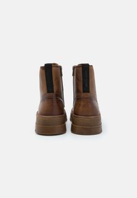Botas de tobillo de cuero marrón con un acabado texturizado, cierre de cremallera expuesto y acentos elásticos negros en los lados; suelas de goma.
