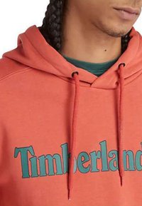 Hoodie corail orange avec le logo "Timberland" vert. Fabriqué en tissu doux, il est doté d'une capuche avec cordon de serrage et d'une poche kangourou, avec des poignets côtelés.