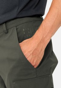 Dunkelgrüne Hose aus leichtem, glattem Material. Verfügt über eine Seiten Tasche und einen sichtbaren Aufdruck mit der Aufschrift "BIOBASIERT." Hand in der Tasche.