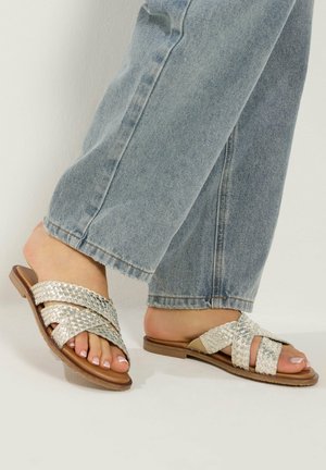 Jalat, joissa on metallinhohtoiset punotut liukusandaalit, yhdistetty vaaleansinisiin farkkuihin, seisomassa vaalealla pinnalla.