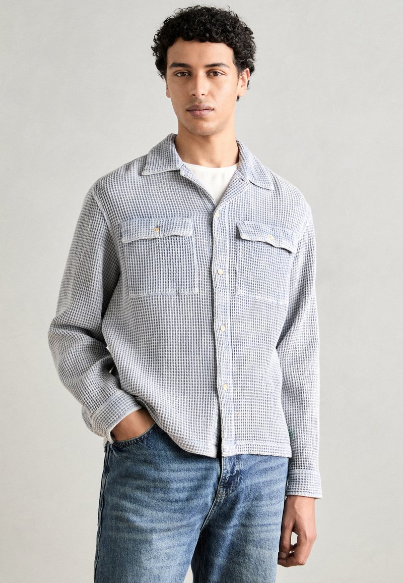 Blå og hvit rutete skjorte med kragehals, to frontlommer, knapper og en strukturert tekstur. Brukt med denimjeans.