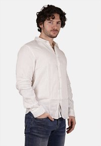 Camicia bianca in lino a maniche lunghe con chiusura frontale a bottoni, colletto alla cinese e tessuto testurizzato, abbinata a jeans in denim blu.