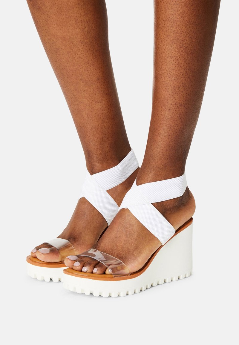 Sandales compensées blanches avec brides transparentes et bandes de cheville en tissu texturé. La semelle est épaisse avec un motif rainuré pour une meilleure adhérence.