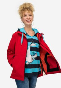 Giacca impermeabile rossa con zip frontale, tasche laterali e cappuccio regolabile, indossata sopra una maglietta a righe blu con stampa grafica.