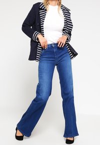 Kvinna klädd i marinblå randig jacka, vit topp, blå hög midja vida jeans och svarta klackar, står med benen korsade mot vit bakgrund.