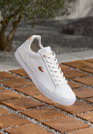 White sneaker featuring a smooth synthetic upper, lace-up design, rose gold accents, and textured rubber sole, displayed on a wooden surface.