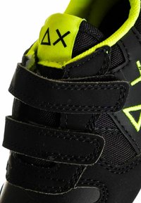 Sneaker neri con cinturini in velcro, dotati di una tomaia in tessuto texturizzato e accenti gialli brillanti sul colletto e sul logo.