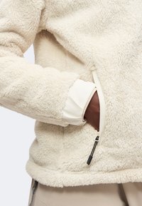 Άτομο με μπεζ fleece μπουφάν με το χέρι μέσα στην τσέπη με φερμουάρ, δείχνοντας μαλακή υφή και στρωμένα λευκά και καφέ μανίκια.