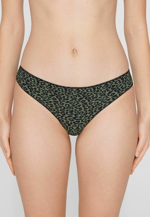 Frau trägt grüne und schwarze Leopardenmuster-Bikini-Unterwäsche und steht mit den Händen seitlich vor neutralem Hintergrund.