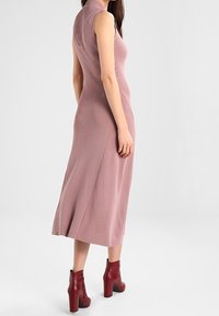 Donna che indossa un abito midi senza maniche color malva e stivali alla caviglia rossi con tacco alto, in piedi con il corpo leggermente girato di lato.