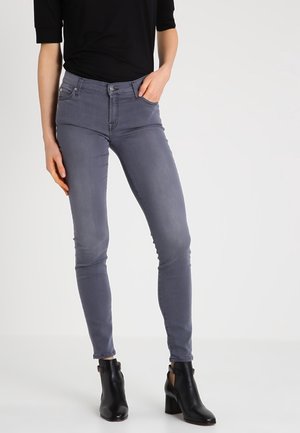 Frau trägt enganliegende graue Jeans, schwarzes Oberteil und schwarze Stiefeletten mit Blockabsatz, steht vor einem schlichten hellen Hintergrund.