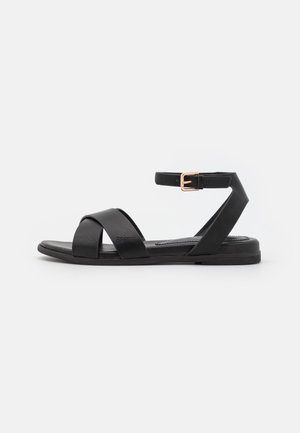 Sandale plate noire avec des brides croisées à l'avant et une bride de cheville avec boucle dorée, présentée sur fond blanc.