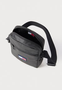 Kompakti musta olkalaukku säädettävällä remmillä, etuosan vetoketjutaskulla ja punaisella, valkoisella ja sinisellä logotarralla etupaneelissa.