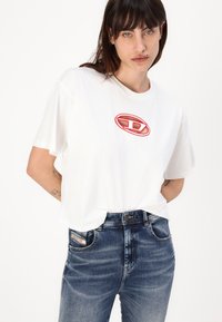 Bijela pamučna majica s crveno-bijelim logotipom na prsima. Nosena s plavim trapericama svijetle pranje s vidljivim šavovima na džepovima.