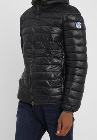 Svart puffjacka med horisontellt quiltat mönster, glänsande textur, dragkedja och huva. Har en cirkulär logotyp på vänster ärm.