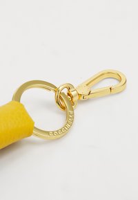 Portachiavi in pelle gialla con hardware dorato, dotato di anello rotondo e moschettone, inciso con "COCCINELLE." Texture liscia.