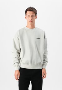 Sweatshirt cinza claro com decote redondo, ombros caídos e punhos canelados; apresenta um logotipo preto "PEGADOR" no peito.