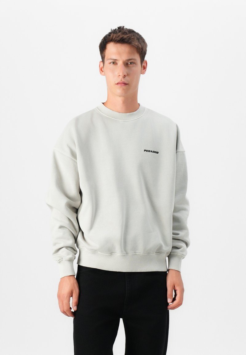 Sweatshirt cinza claro com decote redondo, ombros caídos e punhos canelados; apresenta um logotipo preto "PEGADOR" no peito.