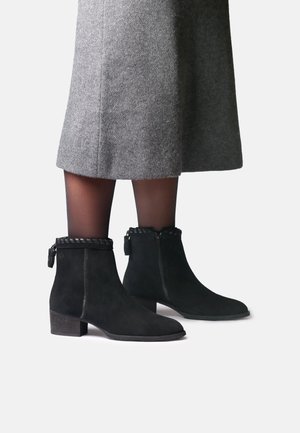Bottines en daim noir à talons blocs bas et garniture tressée portées avec des collants noirs transparents et une jupe grise longueur genou.