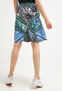 Jupe plissée colorée présentant un mélange de motifs géométriques et floraux en vert, bleu et noir, fabriquée en tissu léger avec une texture lisse.