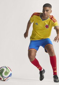 Gul fodboldtrøje med røde og blå accenter, blå shorts, røde sokker og sorte støvler. Bolden har et flerfarvet design.