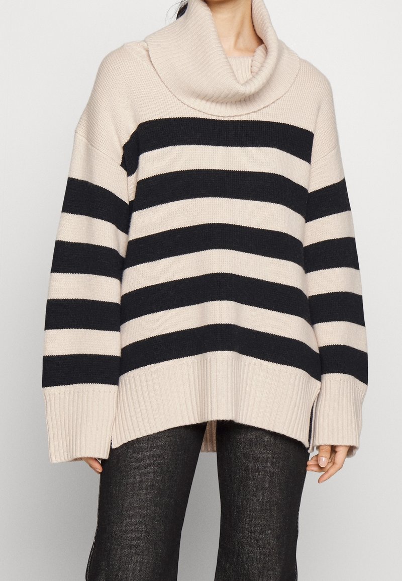 Femme portant un pull beige oversize avec de larges rayures horizontales noires et un grand col roulé plié, associé à un pantalon foncé.