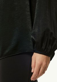 Schwarzes Langarmoberteil mit weicher Textur, das über Puffärmel verfügt und einen Saum hat, der an einer enganliegenden schwarzen Unterlage anschließt.