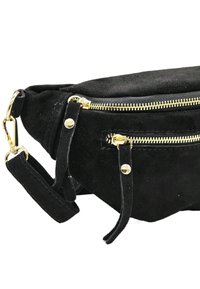 Sac en daim noir avec deux zippers en métal doré, accents en cuir, et une bandoulière détachable. La surface texturée ajoute de la profondeur.