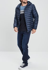 Marineblaue Pufferjacke mit Kapuze, Rippbündchen, getragen über einem blauen gestreiften Pullover und dunklen Jeans, kombinierte mit marineblauen Sneakern.