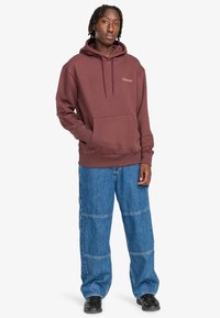 Felpa maroon con tasca e lacci, abbinata a jeans a gamba larga blu con dettagli di cucitura e vestibilità rilassata. Scarpe nere completano il look.