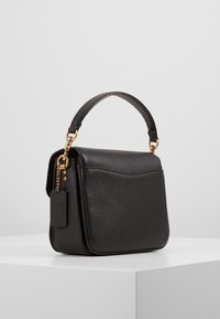 Borsa a mano in pelle nera con forma strutturata, manico superiore, accessori in metallo dorato, superficie texture e dettaglio del tavolo per il bagaglio.