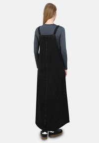 Completo in denim nero con bretelle regolabili, design lungo e fluido, e orlo grezzo, indossato sopra una maglietta grigia a maniche lunghe.