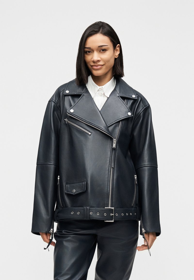 Νεαρή γυναίκα με ίσια μαύρα μαλλιά που φοράει μια μαύρη δερμάτινη μπουφάν μοτοσικλετιστή με ζώνη πάνω από ένα λευκό πουκάμισο με γιακά.