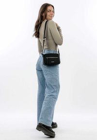 Black crossbody bag with textured leather, front zip pocket, adjustable strap, and rectangular shape. Worn over light blue denim jeans.