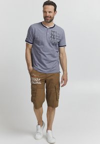 Jūrinės ir pilkos spalvos henley marškiniai su baltais akcentais, turintys margintą tekstą. Rudi kroviniai šortai su šoninėmis kišenėmis ir grafiniu dizainu. Balti sportbačiai.