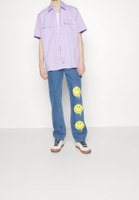 Lavendel kortärmad skjorta med två bröstfickor, tillsammans med blå jeans med tre stora gula smiley-ansikten. Vita sneakers fulländar looken.